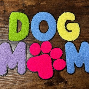 May include: A collection of colourful, textured patches spelling "DOG MOM" with a paw print. The letters are green, blue, yellow, purple, and blue, with a hot pink paw print. Each letter has a black outline.