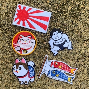 May include: A collection of embroidered patches featuring Japanese cultural symbols. Includes a rising sun flag, a lucky cat, a sumo wrestler, a Shiba Inu dog, and two koi fish wind socks.