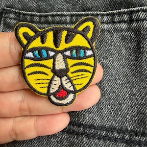 Puede incluir: Un parche bordado que muestra una cara de tigre estilizada con pelaje amarillo brillante, rayas negras, ojos azules penetrantes, un hocico blanco y una pequeña boca roja, delineado con hilo negro sobre una tela de mezclilla gris oscuro.