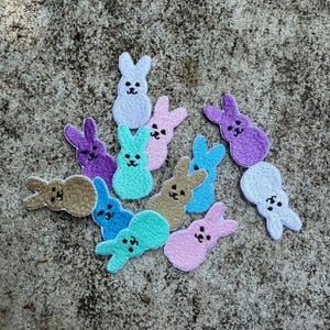 May include: A collection of embroidered bunny patches in various pastel colors, including blue, purple, pink, and green. Each patch features a simple bunny shape with embroidered facial details. These patches are likely for crafting or embellishment.