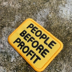 May include: Yellow patch with black text that reads "PEOPLE BEFORE PROFIT." The patch has a yellow border and is set against a textured, gray background. The patch is a square with rounded corners.