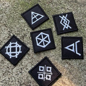 Może przedstawiać: Zestaw sześciu czarnych kwadratowych naszywek z białymi haftowanymi geometrycznymi symbolami. Naszywki są ułożone na teksturowanej, szarej powierzchni. Każda naszywka ma unikalny wzór, w tym trójkąty, linie i okrągłe wzory.