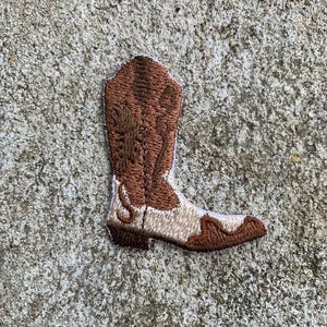 Könnte beinhalten: Gestickter Aufnäher in Form eines Cowboy-Stiefels. Der Stiefel hat braune und beige Farben mit detaillierten Nähten. Der Aufnäher ist zum Aufbügeln oder Aufnähen auf Kleidung oder Accessoires gedacht.