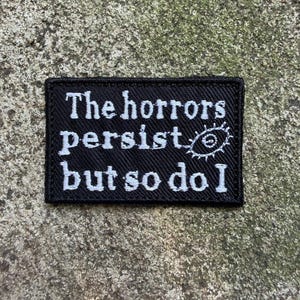 May include: Black rectangular patch with white embroidered text that reads "The horrors persist but so do I." A stylized eye is embroidered to the right of the text. The patch is on a textured, gray surface.
