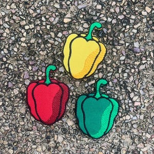 Puede incluir: Tres parches bordados de pimientos en amarillo, rojo y verde, cada uno con un tallo verde. Los parches están colocados sobre una superficie de piedra texturizada, gris y marrón. Los pimientos están delineados en negro.