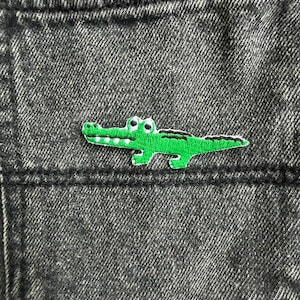 May include: A black acid-wash denim fabric featuring an embroidered patch of a green crocodile. The crocodile is depicted in profile with white teeth, black eyes, and a textured green body, positioned horizontally across the centre of the fabric.