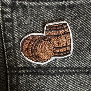 May include: Embroidered patch depicting two brown barrels with a white border, set against a dark grey denim background. The patch features detailed stitching, highlighting the barrel's wooden texture. Suitable for denim jackets or bags.