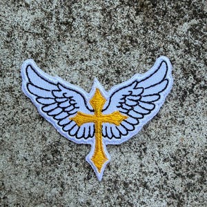 Może przedstawiać: Haftowana naszywka przedstawiająca złoty krzyż z białym obrysem, umieszczony na parze białych skrzydeł z czarnymi detalami. Naszywka znajduje się na teksturowanej, szarej powierzchni. Wzór jest symbolem religijnym.