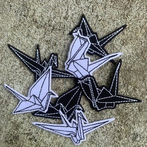 May include: A collection of origami crane patches in black and white. The patches are embroidered with detailed stitching, showcasing the folded paper design. The cranes are arranged in a circular pattern, with some facing different directions.