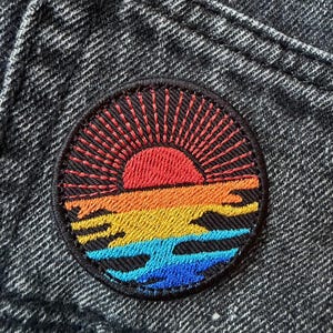 Könnte beinhalten: Runder, gestickter Aufnäher mit einem Sonnenuntergangsmotiv. Die Sonne ist rot mit orangen Strahlen, überlagert von orangefarbenen, gelben und blauen Wellen. Der Aufnäher hat einen schwarzen Rand und ist auf dunklem Denim-Stoff aufgenäht.