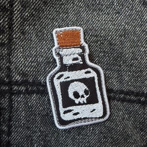 May include: Embroidered patch featuring a black bottle with a skull and crossbones design. The patch has a white border and a brown cork. The patch is likely for clothing or accessories.