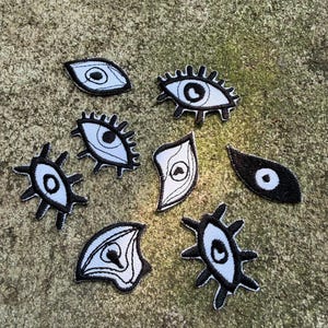 May include: A collection of embroidered eye patches in various shapes and sizes. Each patch features a white eye design with a black outline and details. The patches are set against a textured, neutral-toned background.