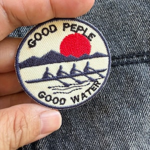 Op de afbeelding: Een ronde geborduurde patch met een crèmekleurige achtergrond, voorzien van een donkerblauw gebergte, een felrode zon en een donkerblauwe roeiboot met vier roeiers. De tekst GOOD PEPLE staat in een boog erboven en GOOD WATER eronder in donkerblauw.