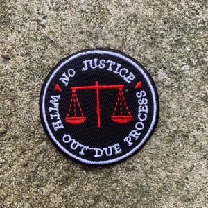 May include: A round, black patch with white text that reads "NO JUSTICE OUT DUE PROCESS". A red scale of justice is in the center. The patch is on a textured, gray surface.