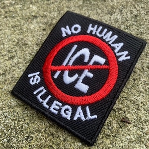 May include: Black square patch with white text and a red circle with a line through it. The text reads "NO HUMAN IS ILLEGAL" with "ICE" in the center of the circle. The patch is on a textured, light brown surface.