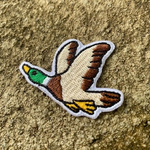May include: Embroidered patch of a flying duck. The duck features a green head, brown and beige wings, and yellow feet. The patch is outlined in white.