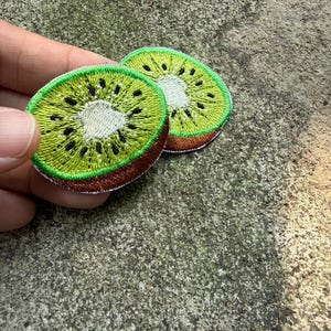 Puede incluir: Dos parches bordados con forma de kiwi. Cada parche presenta una cáscara verde, un borde marrón y un centro blanco con semillas negras. Los parches están diseñados para coser o planchar sobre tela.