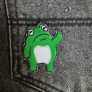 May include: A green frog patch with white belly and black outline is sewn onto a dark gray denim pocket. The frog is waving its right arm. The patch is embroidered with a cartoon-like design.
