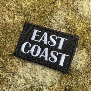 以下が含まれることがあります： 黒い長方形のパッチ。白い大文字で「EAST COAST」と刺繍されています。パッチは、質感のあるオリーブグリーンの背景に配置されています。