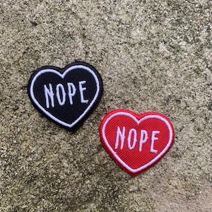 Puede incluir: Dos parches en forma de corazón. Uno es negro con borde blanco y la palabra "NOPE" en blanco. El otro es rojo con borde blanco y la palabra "NOPE" en blanco. Los parches están sobre una superficie gris texturizada.