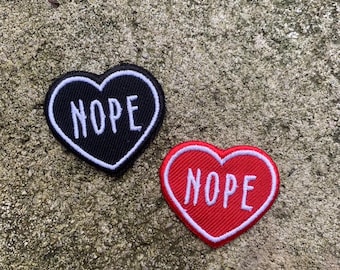 Puede incluir: Dos parches en forma de corazón. Uno es negro con borde blanco y la palabra "NOPE" en blanco. El otro es rojo con borde blanco y la palabra "NOPE" en blanco. Los parches están sobre una superficie gris texturizada.