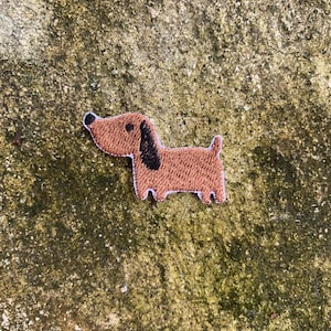 Puede incluir: Un parche bordado marrón de perro salchicha con detalles en negro. El parche presenta un diseño detallado del cuerpo, la cabeza y la cola del perro. El parche está colocado sobre una superficie texturizada, moteada en verde y marrón.