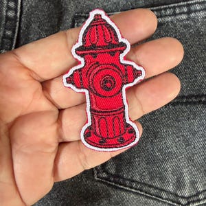May include: A red fire hydrant patch with a white border. The patch is embroidered with black details, including the hydrant's features. The patch is held in a hand against a gray denim background.