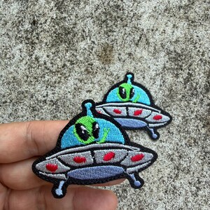 Może przedstawiać: Dwie haftowane naszywki z motywem kreskówkowego UFO. UFO mają jasnoniebieski wierzch, szarą podstawę z czerwonymi akcentami i zielonego kosmitę z czarnymi oczami. Naszywki znajdują się na teksturowanej szarej powierzchni.