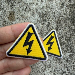 May include: Two yellow triangular patches with a black lightning bolt symbol. The patches have a black border and white stitching. The background is a textured, gray surface.