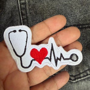 Op de afbeelding: Geborduurde patch met een stethoscoop, een rood hart en een ECG-hartslaglijn. De patch is wit met zwarte en rode details, geschikt voor medische professionals of gezondheidsenthousiastelingen.