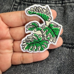 May include: An embroidered patch featuring a cluster of monstera leaves with vibrant green detailing and white borders. The design showcases intricate black stitching that outlines the leaf shapes, creating a textured appearance on a white fabric base.