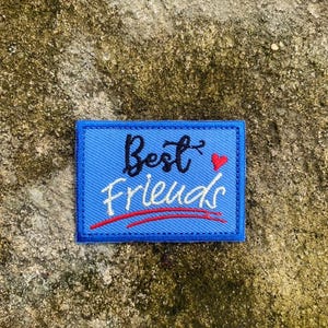 May include: A blue rectangular patch with the words "Best Friends" in black script and white cursive, with a red heart and a red underline. The patch is on a textured, tan surface.