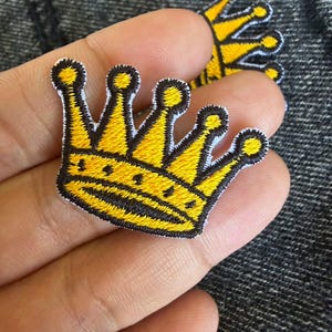 May include: A yellow and black embroidered crown patch featuring five points topped with small circles. The design includes a thick black border and detailed yellow stitching throughout the crown shape, creating a textured appearance for this accessory.