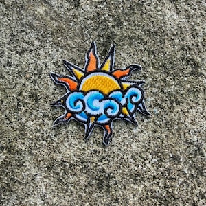 Könnte beinhalten: Gestickter Aufnäher mit einer stilisierten Sonne mit orangefarbenen und gelben Strahlen, einem gelben Zentrum und blau-weißen Wolken. Der Aufnäher hat einen schwarzen Rand und ist für Kleidung oder Accessoires geeignet.