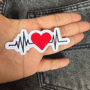 Peut inclure: Écusson brodé avec un cœur rouge au centre, intégré dans un motif d'électrocardiogramme noir et blanc. Le patch a une bordure blanche et est destiné aux vêtements ou accessoires.