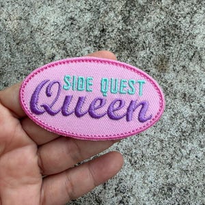 May include: An oval-shaped embroidered patch in pink, featuring the text "SIDE QUEST Queen" in purple and teal. The patch has a pink border and is designed to be attached to clothing or accessories, possibly by sewing or ironing.