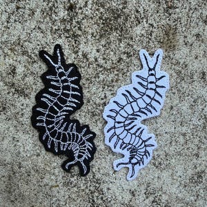 Puede incluir: Dos parches bordados con forma de ciempiés. Uno es negro con detalles blancos, el otro blanco con detalles negros. Los parches están colocados sobre una superficie gris texturizada.