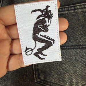 May include: A white rectangular patch with a black illustration of a devil-like figure. The figure is kneeling with a tail and horns. The patch is held against a denim background.