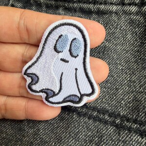 Può includere: Una toppa ricamata raffigurante un fantasma bianco con occhi ovali azzurri e una piccola linea orizzontale per la bocca. Il fantasma presenta un contorno nero cucito e delicate sfumature azzurre lungo i bordi inferiori della sua forma.