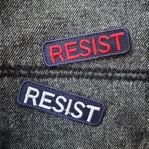 May include: Two rectangular embroidered patches with the word "RESIST". One patch has red text on a navy blue background, the other has white text on a navy blue background. The patches are sewn onto a dark denim fabric.