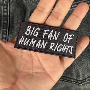 Peut inclure: Un écusson rectangulaire noir avec le texte brodé en blanc "BIG FAN OF HUMAN RIGHTS". L'écusson est tenu dans une main sur un fond en denim foncé.