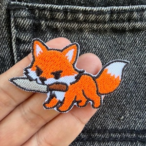 Pode incluir: Um emblema bordado apresentando uma raposa estilizada laranja e branca com um contorno preto. A raposa é retratada de perfil, segurando uma faca prateada com cabo marrom na boca. O design inclui detalhes brancos nas orelhas, no rosto e na cauda.