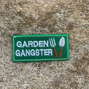 May include: A green rectangular patch with the words "GARDEN GANGSTER" in white, embroidered text. The patch features a brown-handled garden fork and trowel. The patch is likely for clothing or accessories.