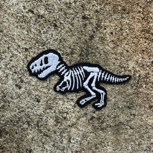 Könnte beinhalten: Ein schwarz-weißer, gestickter Aufnäher mit einem Dinosaurierskelett-Design. Der Aufnäher zeigt ein Tyrannosaurus Rex-Skelett in einer Laufpose. Der Aufnäher befindet sich auf einer strukturierten, grauen Oberfläche.