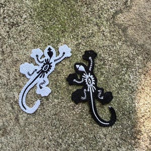 May include: Two embroidered lizard patches. One is black with white detailing, the other is white with black detailing. Both have a detailed design on the body and a curved tail. The patches are on a textured, grey surface.