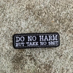 Peut inclure: Un écusson rectangulaire noir avec du texte blanc brodé. Le texte dit "DO NO HARM BUT TAKE NO SHIT". L'écusson est conçu pour être cousu ou repassé sur des vêtements ou des accessoires. Le fond est une surface grise texturée.