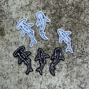 Peut inclure: Six écussons brodés représentant des requins. Trois écussons sont blancs avec des contours noirs, représentant des requins marteaux et d'autres espèces. Les trois autres écussons sont noirs avec des contours blancs, représentant des silhouettes de requins.