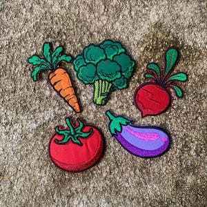 May include: A set of embroidered vegetable patches. The collection includes a carrot, broccoli, beetroot, tomato, and aubergine. Each patch is brightly coloured with a detailed design and stitched border, suitable for customising clothing or accessories.