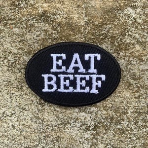 Könnte beinhalten: Schwarzer ovaler Aufnäher mit dem weiß gestickten Text "EAT BEEF". Der Aufnäher ist wahrscheinlich für Kleidung oder Accessoires gedacht und wird auf einer strukturierten, hellbraunen Oberfläche gezeigt.