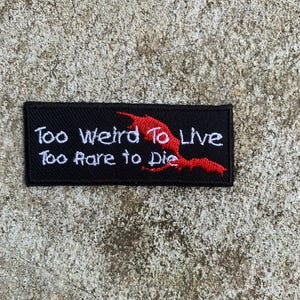 Op de afbeelding: Zwart rechthoekig embleem met witte tekst: "Too Weird To Live, Too Rare to Die." Een rode vleermuisachtige afbeelding staat rechts. Het embleem ligt op een getextureerd grijs oppervlak.